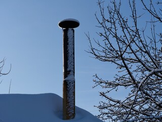 A snow-covered roof with old metal chimney and tree branches against blue sky after snowfall.