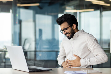 A young Indian businessman is suffering from severe stomach pain. Sitting in the office at the desk and holding hands, bent and grimacing in discomfort - obrazy, fototapety, plakaty