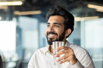 Close-up photo of a young man, an Indian businessman sitting in the office and holding a glass of clean water in his hands, quenching his thirst, smilingly looking to the side