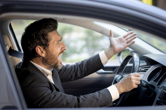 Close-up photo of angry young businessman in suit driving car and emotionally gesturing with hands, displeased shouting and arguing