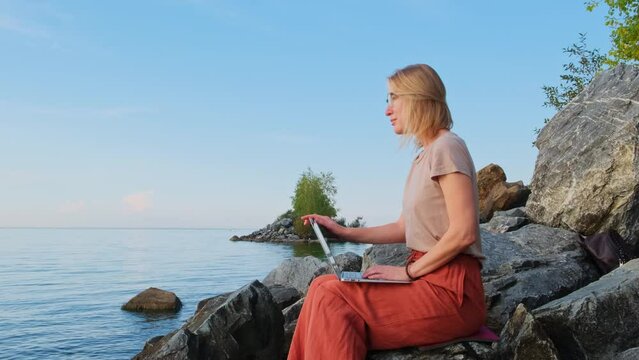 A digital nomad, blonde caucasian woman sits by the sea and works remotely. Female freelancer uses laptop computer on empty seashore. Office for introvert outdoor.