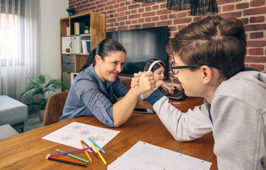 A mother playfully arm wrestles with her son at a wooden table while her daughter is busy on a laptop in the background