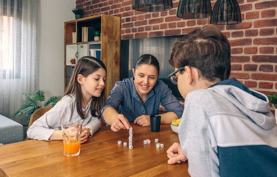 A mother with her children smiling and playing a dice game at the family table with snacks and drinks during a cozy weekend at home