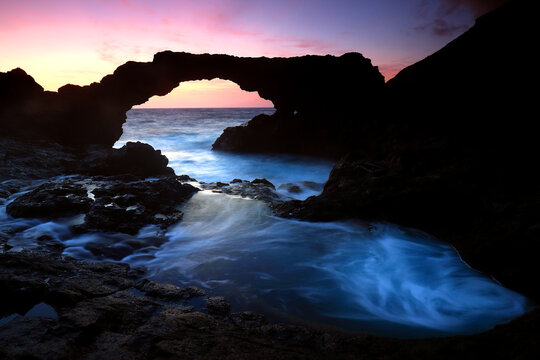 A serene dawn breaks over El Hierro, casting a soft glow through a natural volcanic rock arch over swirling sea waters