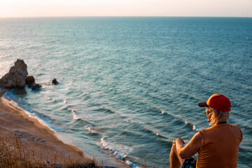 mature man sits on top of a mountain on the seashore and looks at the sunset.Travel and tourism.