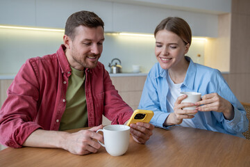 Cheerful happy woman and glad smiling man spend free time, weekend enjoying tea, coffee drinking, surfing in internet, scrolling social networks. Independent living, joint residence, marriage concept