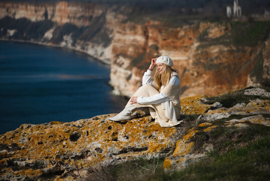 young blonde woman 35 - 40 years old in white clothes, long sweater, boots, beret sits on the ground against the backdrop of Cape Emine in Bulgaria and looks towards the sea smiling, spring, autumn
