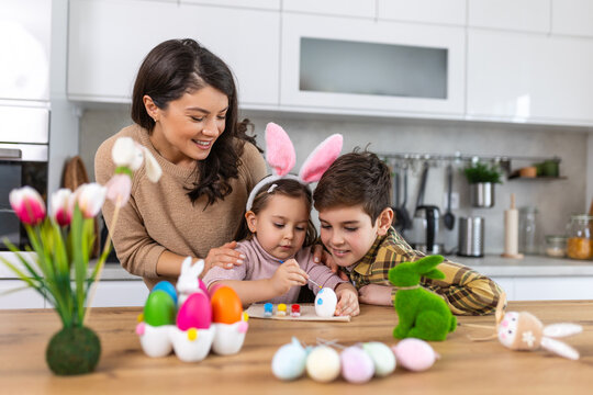 Cute little children wearing funny bunny ears headbands embracing and kissing young happy mother while painting Easter eggs together, sitting at table in kitchen, selective focus