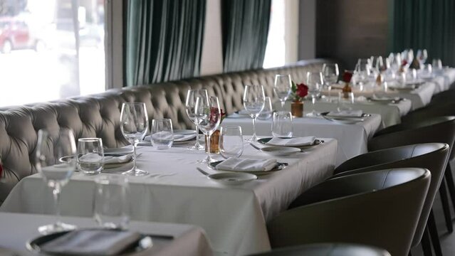 Well-set dining table in restaurant beside window with natural light. Plush leather sofa running along length of desk. Small vases with red roses and wine glasses