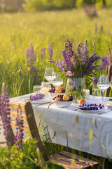 Elegant gorgeous wedding table decor or romantic dinner arrangement outdoors in blooming field. Purple lupine flowers, candles, fruits and wine, wooden vintage furniture. Sunset, summer, golden hour