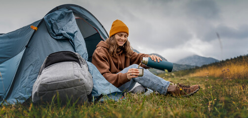 A young cheerful woman traveling with a tent pouring hot tea from a thermos. Hiking and trekking concept - obrazy, fototapety, plakaty