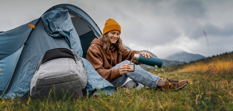 A young cheerful woman traveling with a tent pouring hot tea from a thermos. Hiking and trekking concept