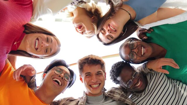 Low angle view of a group of multiracial friends standing on a circle, smiling and embracing together. Young teenagets laughing and looking at camera. Team of confident people on a coaching meeting