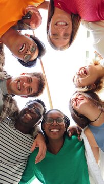 Vertical low angle view of a group of multiracial friends standing on a circle, smiling and embracing together. Young teenagers laughing and looking at camera. Team of people on a coaching meeting