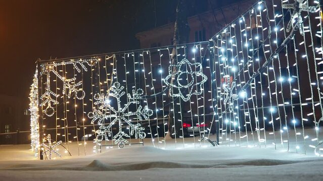 Glowing snowflakes. Festive New Year Christmas electric art installation architecture structures in snowdrift. Light decoration of city street. Winter night. Garland light bulbs shine in silver. Xmas