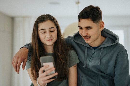 Brother with arm around sister using smart phone at home