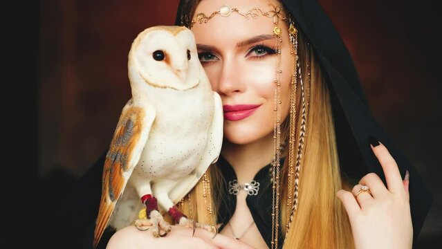 Art portrait fantasy gothic mystical woman holding white barn owl in hand, hiding part of face. Fantasy Elf Blonde hair sorceress girl sexy eyes lips looking into camera Wild bird wings. wizard hood