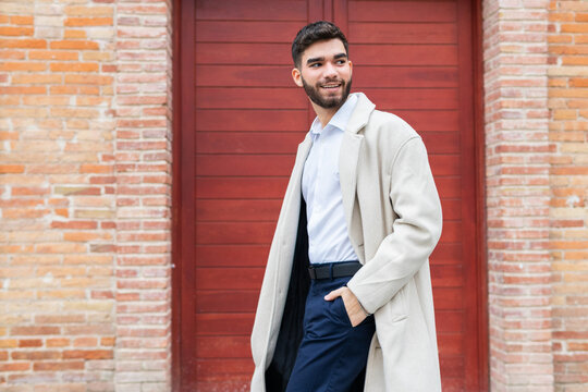 A stylish young man in business attire smiles confidently while standing against a red door backdrop, exuding a sense of casual professionalism in his beige coat and navy trousers