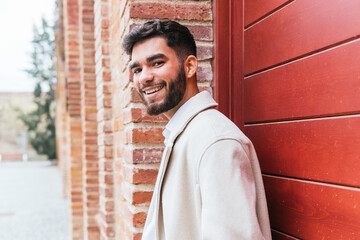 Charming businessman in a stylish coat smiling and leaning against a red brick wall, radiating confidence and ease