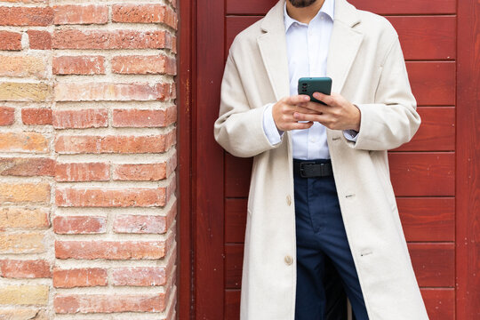 Close-up of anonymous businessman in a trench coat using a smartphone against a textured brick wall, embodying urban professionalism