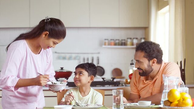 Happy Indian woman serving food to her son and husband on dining table at home - concept of family bonding, household responsibility and togetherness