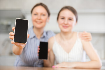 Cheery middle-aged mother and young daughter demonstrating their phones sitting at table in the kitchen