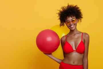 African American woman in red swimsuit having fun holding inflatable ball and going on summer holiday trip standing on yellow studio background. Vacation tour and travel concept.