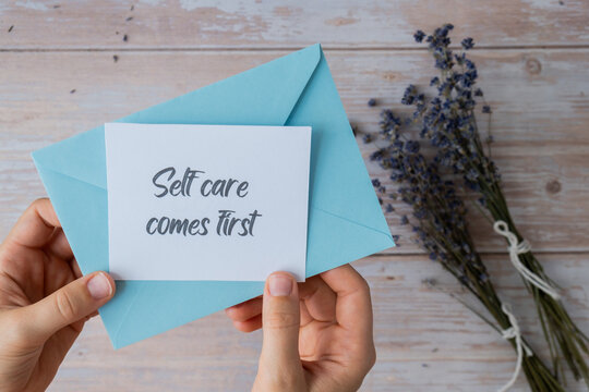 Female hands taking paper card note with text SELF CARE COMES FIRST from blue envelope. Lavender flower. Top view, flat lay. Concept of mental spiritual health self care wellbeing