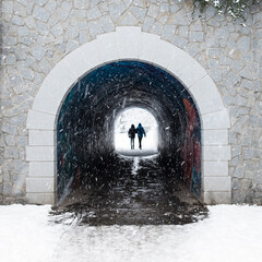 A silhoette of a romantic couple holding hands on a winter walk for the Valetines dinner walking through a dark pedestrian tunnel to white snowy garden. Light at the end of  the tunnel. 