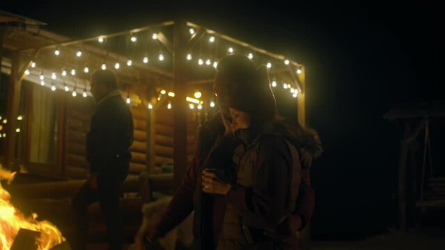Multiethnic couple of travelers drinks tea, laughs and warms standing near the fire outdoors during journey. In the background tourist friends rest in stylish wooden gazebo decorated with light bulbs.