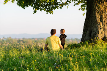 Dad with son in the spring meadow sitting under the tree in tall grass. Father and son outdoors. Travel with child concept