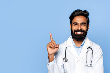 Smiling middle aged indian male doctor pointing up on blue backdrop - obrazy, fototapety, plakaty