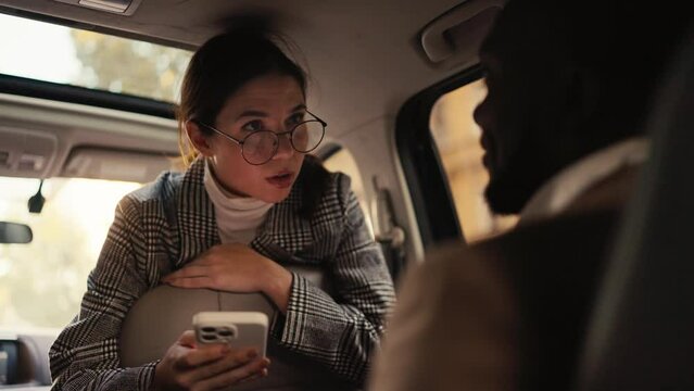 A confident brunette girl in round glasses in a gray jacket communicates with a man with Black skin in a brown jacket during her business meeting and business trip in a modern car interior