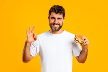 Portrait of cheerful man gesturing okay sign holding takeaway burger