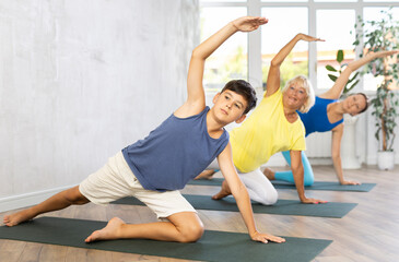 Diligent preteen boy practicing half side plank pose of yoga on black rug in light fitness room
