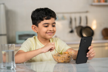 Indian preteen kid eating noodles while using mobile phone at home on dining table - concept of...