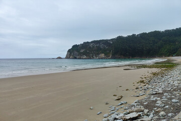 A serene beach with calm waters, rocky formations, and greenery under an overcast sky, evoking a tranquil atmosphere