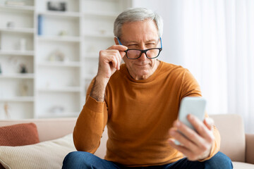 Elderly man adjusting his eyeglasses while using smartphone at home