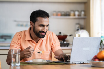 Indian middle aged man working on laptop while eating food on dining table at home - concept of...