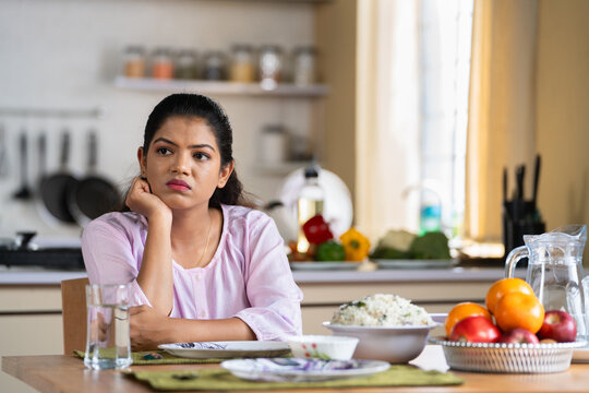 Indian woman on dining table waiting for husband or child after preparing food at home for lunch - concept of family caring, responsibility and patience