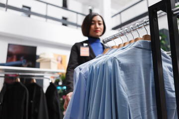 Blue casual shirts hanging on apparel rack while asian woman seller standing in mall clothing store...