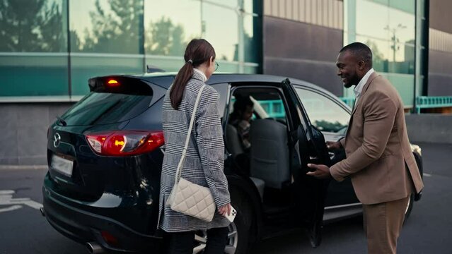 Confident and happy man Businessman with Black skin and a beard in a brown suit invites a brunette female colleague to sit in the passenger seat of a black car and sits behind her during their