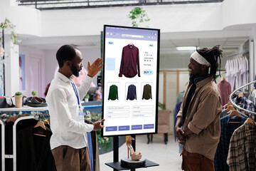 Young worker showing items on touch screen monitor kiosk service to client in fashion boutique at...