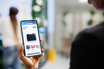 Client choosing clothes on online app, looking at modern fashion collection on mobile application. Young shopper choosing clothing items on clothes boutique website in retail shop.