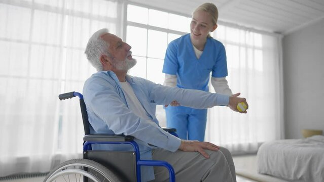Man in wheelchair exercising with nurse. Social medical worker female helping senior male doing physical hands exercises with dumbbell. Palliative care in late stage of neurodegenerative pathologies.