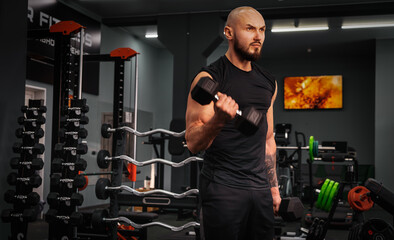 An athlete with a beard performs an exercise with dumbbells in an equipped bodybuilding gym.