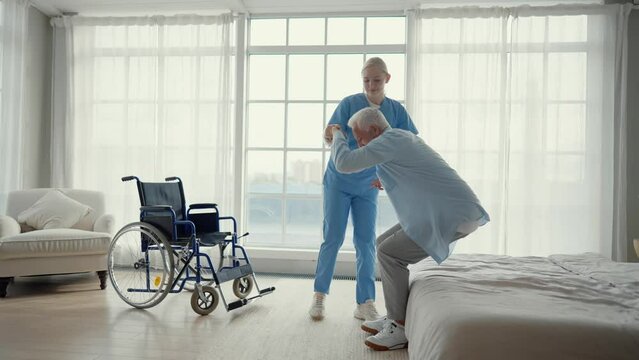 Nurse helps old wheelchair man to stand up. Physical therapist woman provides medical help support to senior paralyzed male standing walking. Care in late stage of neurodegenerative pathologies.