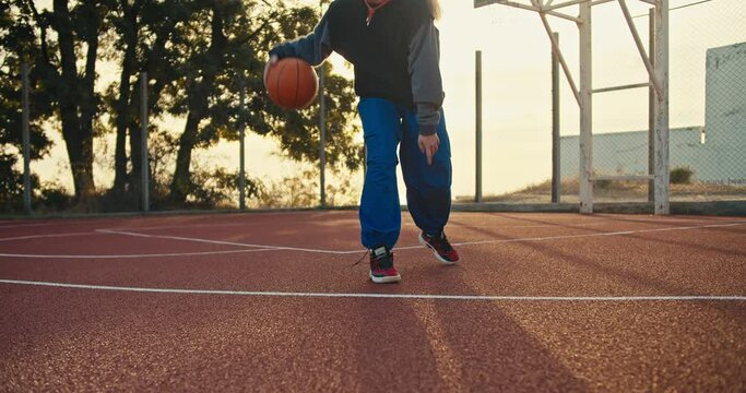 A blonde girl in a sports uniform maneuvers with the ball bouncing it off the floor during her game of basketball on the Red street court in the summer morning
