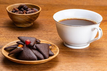 High angle view of delicious homemade crispy dry mixed fruit brownie crackers in wooden plate with cacao nibs in wooden cup, and a white ceramic cup of coffee on wooden table.