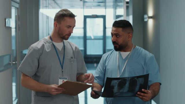Two male doctors in medical uniforms discussing x-ray scan while walking together through corridor in clinic